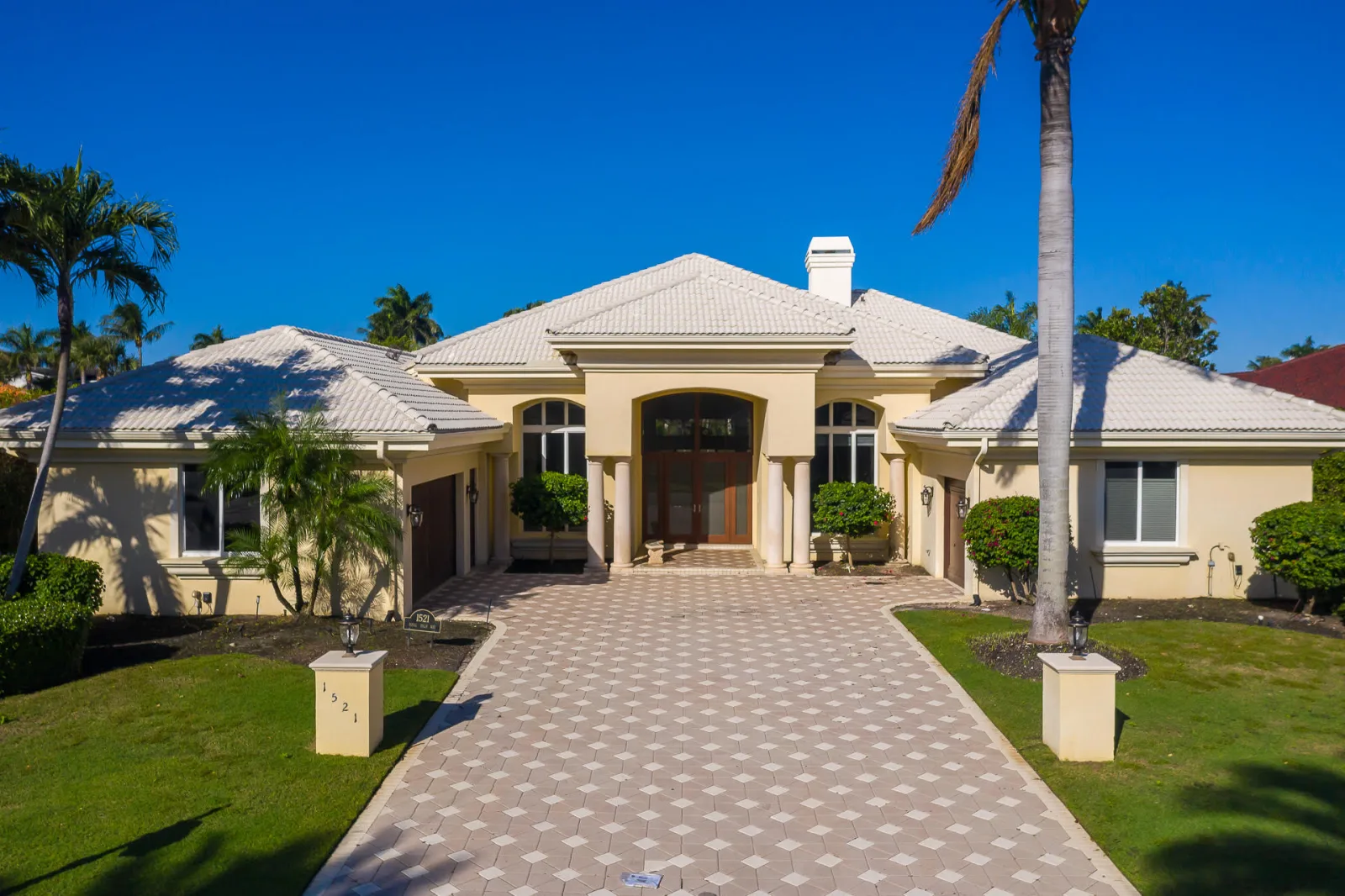The front elevation of 1521 Royal Palm Way, four round columns framing a deep arched entrance beneath a pale tile roof, with two-tone diamond paving sweeping to the garage