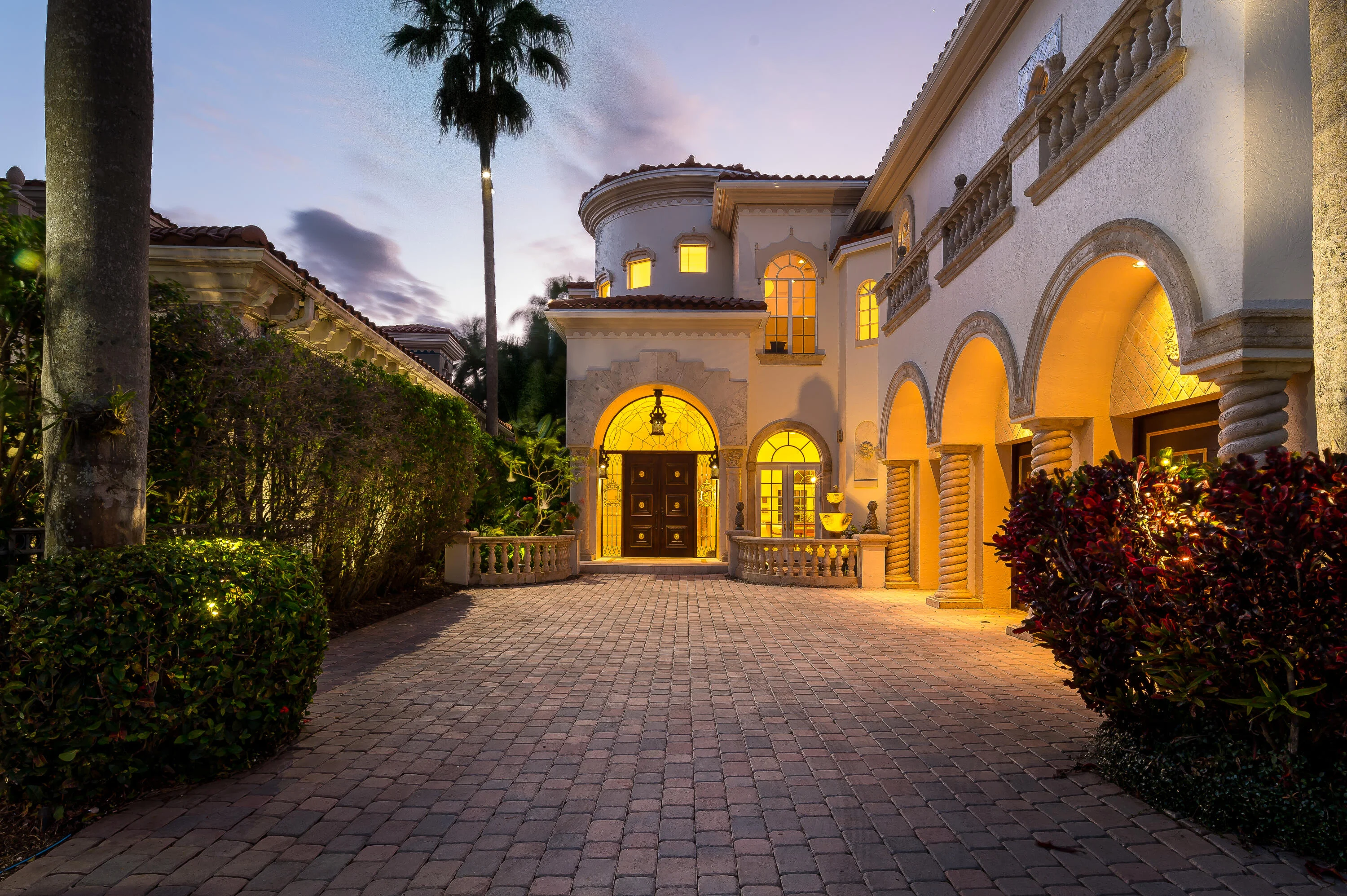 422 Addison Park Lane at dusk, the paver motor court leading to the lit arched entrance beneath the round turret and the second-floor loggia