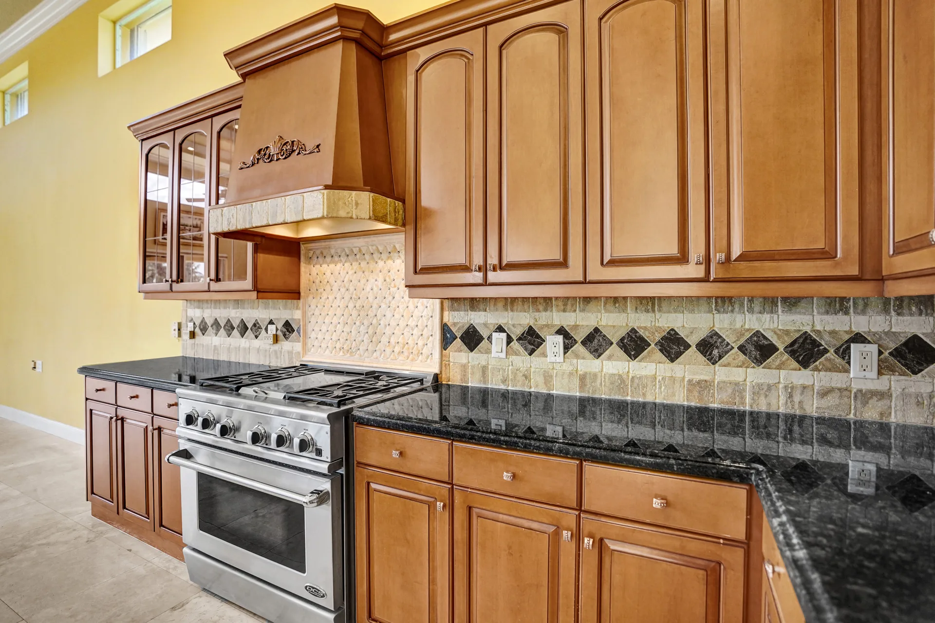The cooking wall at 6543 Newport Lake Circle, a carved wood hood above tumbled-stone tile set with dark diamond inserts and a basket-weave panel behind the range