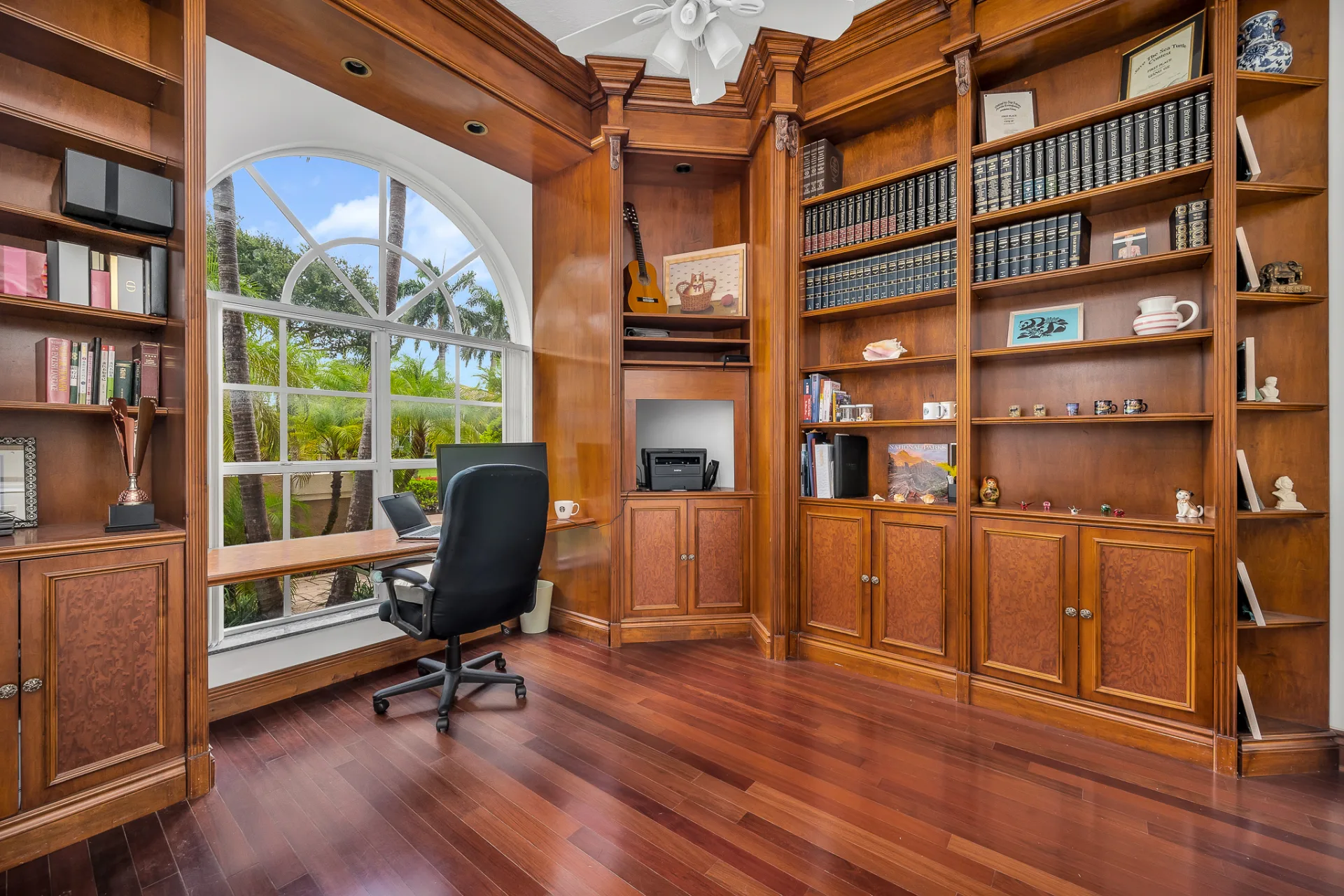 The library at 6662 Newport Lake Circle, wall-to-wall shelving in warm wood above burl cabinet fronts, with a built-in desk beneath a tall arched window whose white muntins radiate in a fan
