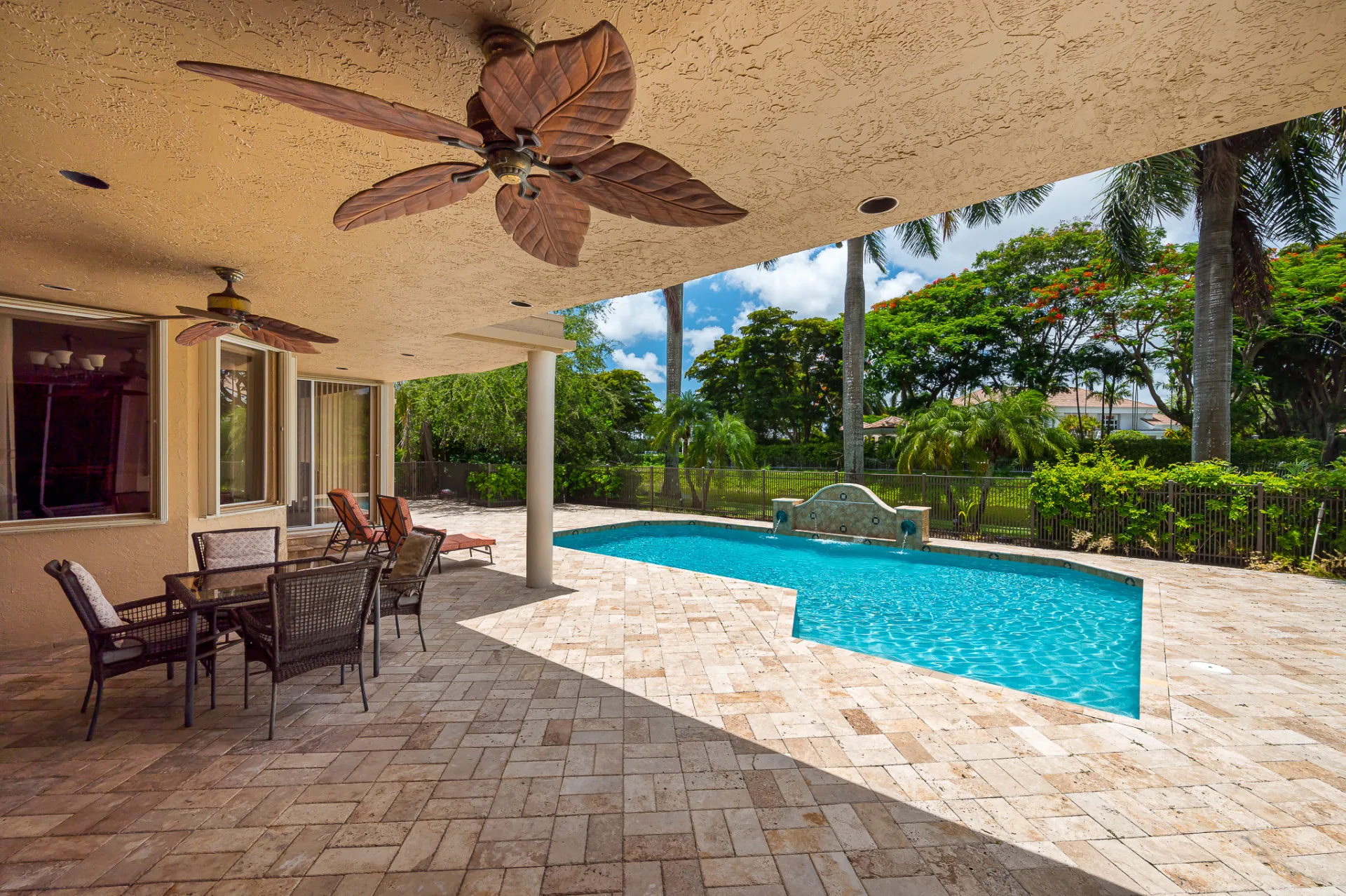 The covered patio at 6662 Newport Lake Circle, palm-leaf ceiling fans above sheltered dining, with steps down to the paver deck and the pool beyond