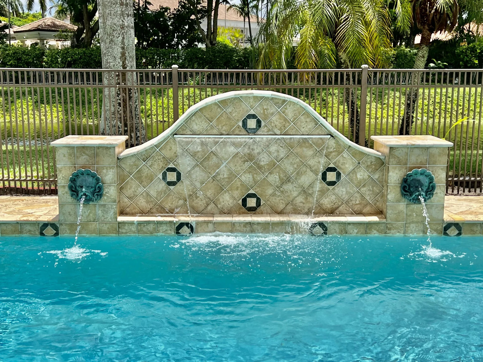 The fountain wall at 6662 Newport Lake Circle, two green-patina lion masks arcing water toward the pool while two further sheets descend across the diamond-patterned centre